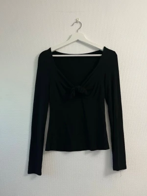 Svart långärmad topp med knyt - Snygg svart långärmad topp med v-ringning och knyt framtill. Figurnära passform och mjukt, stretchigt material som känns bekvämt. Perfekt att styla med jeans eller kjol för en trendig look.