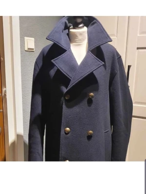 Mörkblå dubbelknäppt rock - Stilren mörkblå rock med dubbelknäppning och stora slag. Klassisk modell med fyra dekorativa metallknappar framtill och två sidofickor. Tillverkad i ullmaterial som ger en snygg och tidlös look. Perfekt för dig som vill ha en rock med lite edge.