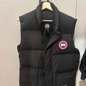 Svart dunväst från Canada Goose - Kvitto och orginal tags från johnells följer självklart med! Svart dunväst från Canada Goose med klassisk röd och vit logotyp på bröstet. Västen har hög krage, två stora fickor framtill och stängs med både dragkedja och tryckknappar. Perfekt för kalla dagar och har en stilren, tidlös look. Hann endast använda den en gång förra året! Nyskick!  