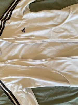 Vit Adidas hoodie med dragkedja - Säljer en vit hoodie från Adidas med dragkedja framtill och klassiska svarta ränder längs ärmarna. Hoodien har huva med dragsko, två fickor och Adidas-logga på bröstet. Tillverkad i mjukt material, perfekt för en sportig och avslappnad stil. Det är verkligen inte samma storlek som det står på tröjan jag skulle säga att det är s-xs.