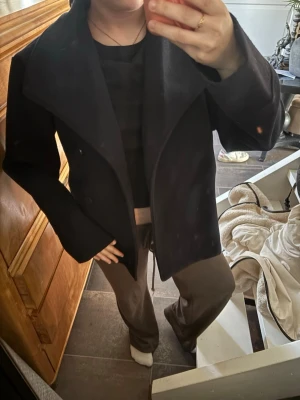 Svart ullrock med bred krage - Stilren svart rock med bred, markerad krage och dubbelknäppning framtill. Rocken har en rak passform och är tillverkad i ett tjockare ullmaterial som ger en snygg siluett. Perfekt för dig som vill ha en klassisk men modern look.