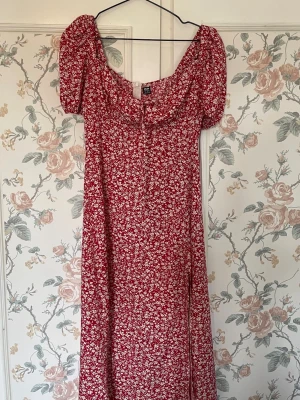 Röd blommig långklänning med puffärm - Långklänning i rött med små vita blommor, puffärmar och söt knytning framtill. Klänningen har en feminin siluett och är perfekt för sommarens alla tillfällen. Tillverkad i ett lätt och luftigt material.