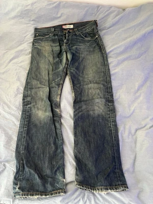 Levi's 506 Standard blå jeans - Klassiska blå Levi's 506 Standard jeans med rak passform och slitna detaljer. Jeansen har fem fickor, orange sömmar och är tillverkade i robust denim. Perfekta för en avslappnad och tidlös stil.