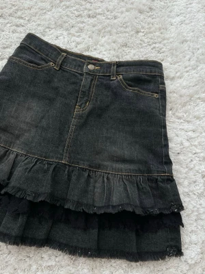 Svart jeanskjol med volangdetaljer - Säljer en svart jeanskjol med dubbla volangdetaljer och råa kanter. Kjolen har klassiska fickor fram och bak samt synliga sömmar i kontrastfärg. Perfekt för dig som gillar en edgy och trendig stil. Står storlek 150 men det är en xs/s
