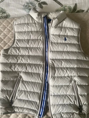 Ljusgrå dunväst från Polo Ralph Lauren - Snygg ljusgrå dunväst från Polo Ralph Lauren med blå logga på bröstet. Västen har två dragkedjeförsedda fickor och en ståkrage. Fodrad med horisontella sömmar och cool randig detalj längs dragkedjan. Perfekt för lager på lager och streetwear.  Storlek L14-16(barnstorlek).
