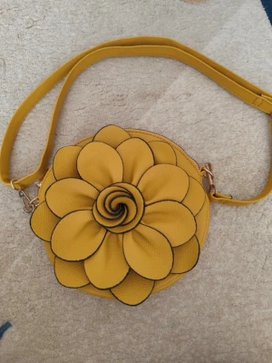 Gul axelväska med blomdetalj - Säljer en rund axelväska i gul konstläder med en stor blomma på framsidan. Väskan har justerbar axelrem och en liten dragkedjeficka på baksidan. Perfekt för dig som vill sticka ut med en unik accessoar.