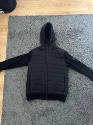 Svart hoodie från J.Lindeberg - Snygg svart cardigan från J.Lindeberg med quiltad front och mjuka ärmar. Dragkedja framtill, två fickor och ribbade muddar. Märkets logga på ärmen ger en clean och modern look. Perfekt för dig som gillar stilrena och sportiga plagg. Lite defekt ner till men inget som syns då det är på insidan 