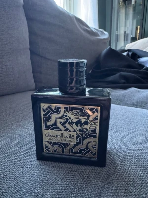 Qaed Al Fursan parfym svart flaska - Snygg parfym från Lattafa i en svart, fyrkantig flaska med ett detaljerat silvermönster och en häst på framsidan. Flaskan har en robust, rund kork och ger ett lyxigt intryck. Perfekt för dig som gillar exklusiva dofter och stilren design.