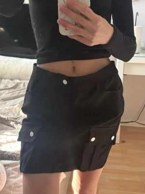 Svart kortkjol med fickor och knappar - Svart kjol med fickor och silverknappar. Aldrig använd med prislapp kvar. För stor för mig som är XS men hade suttit superfint på S eller M