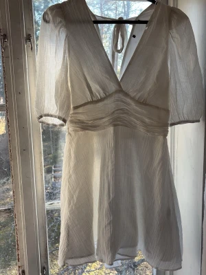 Studentklänning - Säljer en vit kort klänning,djup v-ringning. Klänningen har puffärmar, markerad midja och knytband i nacken. Materialet är tunt och luftigt, perfekt för varma dagar. Har en pytteliten fläck på baksidan (se bild 3)