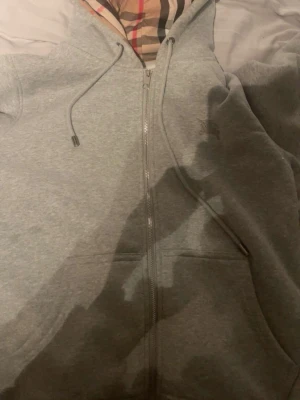 Grå zip hoodie från Burberry - Snygg grå hoodie från Burberry med dragkedja och klassiskt rutigt foder i huvan. Hoodien har dragsko i huvan och två fickor framtill. Perfekt för dig som gillar stilrena plagg med exklusiva detaljer. Materialet är mjukt och bekvämt, troligen bomullsblandning.