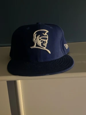 Blå New Era keps med broderad logga - Säljer en blå keps från New Era med en broderad vit logga framtill som föreställer en hjälmprydd profil. Kepsen har rak skärm och klassisk snapback-form. Perfekt för dig som vill ha en stilren och sportig accessoar.