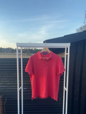 Röd pikétröja från Ralph Lauren - Klassisk röd pikétröja från Ralph Lauren med liten broderad logga på bröstet. Tröjan har krage, knappar vid halsen och korta ärmar. Perfekt för en avslappnad men ändå stilren look.