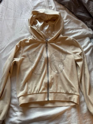 Beige hoodie med fjäril från Lindex - Snygg beige hoodie från Lindex med dragkedja och stor huva. På framsidan finns ett stort fjärilsmotiv i glittriga stenar som ger en cool och unik look. Tillverkad i mjukt material, perfekt för chill dagar eller när du vill sticka ut lite extra.
