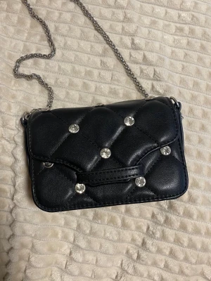Svart quiltad handväska med kedja - Snygg svart quiltad handväska med silverfärgade kedjeremmar och glittrande stenar på både fram- och baksidan. Väskan har en kompakt rektangulär form och är gjord i konstläder. Perfekt för dig som vill ha en trendig accessoar med lite extra bling. Använd få gånger ser helt ny ut.