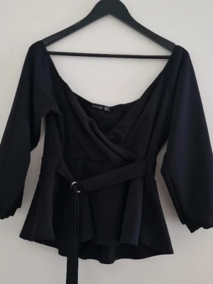 Boohoo Svart off shoulder topp med bälte - Stilren svart omlottblus från Boohoo med v-ringning och markerad midja tack vare det medföljande bältet. Blusen har långa ärmar och ett lätt utställd nederdel som ger en snygg siluett. Perfekt till jeans eller kjol för en trendig look. Perfekt skick. Passar till 44, XXL