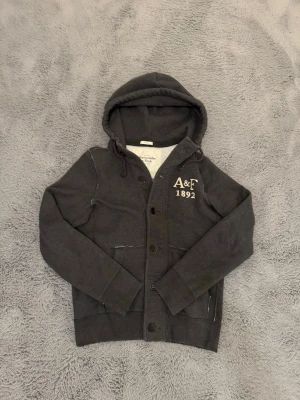 Mörkgrå hoodie från Abercrombie & Fitch - Mörkgrå vintage cardigan med knappar framtill från Abercrombie & Fitch. Broderad logga 'A&F 1892' på bröstet och två fickor fram. Tröjan är i storlek M men passar även S för att den är muskel fit. Tröjan är sjukt sällsynt och eftertraktad då den inte tillverkas längre. Priset är inte hugget i sten så kom gärna med bud!