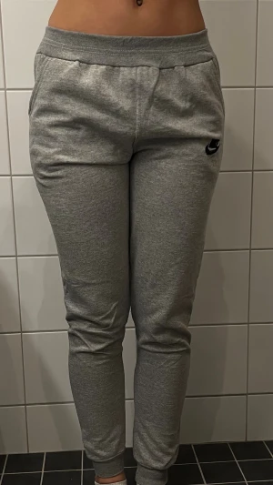 Gråa mjukisbyxor från Nike - Säljer ett par gråa mjukisbyxor från Nike med smal passform och resår vid bensluten. Byxorna har en liten svart Nike-logga på vänster lår och är gjorda i mjukt bomullsmaterial. Perfekta för chill dagar eller träning.