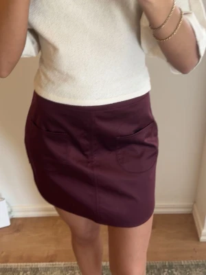 Mörklila kortkjol med fickor - Snygg vinröd kortkjol med två stora fickor framtill. Kjolen har en enkel och clean design som passar perfekt till en casual look. Perfekt för dig som vill ha en stilren och trendig kjol.