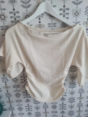 Ljusbeige omlottopp med kort ärm - Trendig ljusbeige topp med omlottdetalj och korta ärmar. Tillverkad i ett strukturerat, lätt material som ger en avslappnad men snygg look. Perfekt att matcha med både jeans och kjol för en stilren outfit.