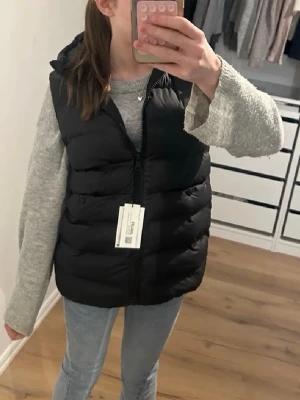 Svart dunväst från Zara - Snygg svart dunväst från Zara med dragkedja framtill. Västen har en puffig design och är tillverkad i ett mjukt, quiltat material. Perfekt att slänga över en hoodie eller tröja för en trendig look. Västen är i storlek 152 men är stor i storleken passar som xs/s.