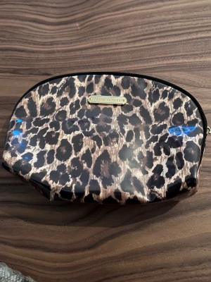 Leopardmönstrad necessär Victoria's Secret - Snygg necessär från Victoria's Secret med leopardmönster i brunt och svart. Väskan har en glansig yta och guldfärgad logga framtill. Perfekt för smink eller småprylar när du är på språng. Praktisk dragkedja upptill och mjuk form som gör den enkel att packa ner.