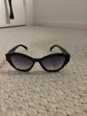 Svarta solglasögon från Prada - Snygga svarta solglasögon från Prada med glansig finish och mörka glas. Klassisk och trendig modell med guldfärgad Prada-logga på skalmen. Säljer åt min mamma till följd av att hon inte längre har någon användning av de. Har bara glasögonen och ingen annan utrustning till den pga att de är använda länge och ”solglasögonväskan” är borttappad, därav det billiga priset.