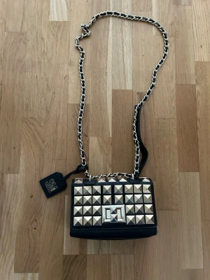Steve Madden Svart axelväska med guldfärgade nitar - Steve Madden. Cool svart axelväska med kedjerem och stora guldfärgade pyramidnitar på framsidan. 