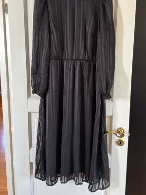 Svart långklänning från Bonprix - Snygg svart långklänning från Bonprix med diskret randigt mönster. Klänningen har långa, transparenta ärmar med resår vid handlederna och en lätt utställd kjol. Perfekt för dig som gillar en elegant och stilren look.