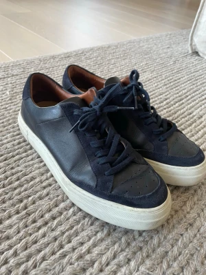 Mörkblå sneakers i läder och mocka J Lindeberg  - Snygga mörkblå sneakers med ovandel i både läder och mocka, vita sulor och mörkblå skosnören. Insidan är fodrad i brunt läder för extra komfort. Perfekta för dig som gillar stilrena och klassiska sneakers.