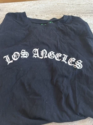 Svart Los Angeles t-shirt Urban Threads - Cool svart t-shirt från Urban Threads med texten 'Los Angeles' i vit gotisk stil på bröstet. Klassisk rund hals och korta ärmar. Perfekt för dig som gillar streetwear och vill ha en enkel men snygg tee i garderoben.