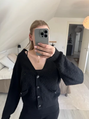 Svart oversized kofta med knappar - Mysig svart oversized kofta med stora knappar framtill och två fickor. Koftan har en djup v-ringning och långa ärmar, perfekt att slänga över en topp. Materialet är mjukt och lite luddigt för extra komfort.