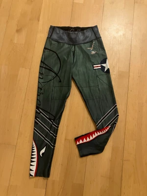 Anarachy apparel Militärgröna leggings med hajmotiv - Säljer ett par militärgröna leggings från Anarchy apparel med hajmotiv och flygplansinspirerade detaljer. Tight passform, elastisk midja och stretchigt material, troligen polyester eller liknande. Snygga vita och röda tänder längs benen och coola tryck som gör dem riktigt unika. Snöret i midajan borta men behövs ej 