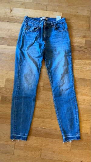 Skinny jeans  - Säljer ett par blå skinny jeans med hög midja och råa, fransiga benslut. Jeansen har klassiska fem fickor och slitna detaljer på bakfickorna. Tillverkade i stretchigt denim för en bekväm passform. Perfekta för en avslappnad och trendig look.