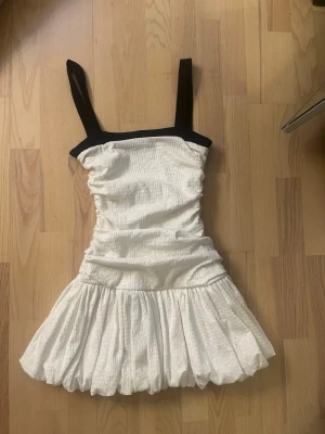 Vit klänning med svarta axelband Zara - Supersöt vit klänning med breda svarta axelband och smockad överdel. Kjolen är voluminös och har en ballongliknande form. Materialet ser ut att vara ett strukturerat tyg som ger en cool och trendig look. Perfekt för dig som gillar kontraster och vill sticka ut.