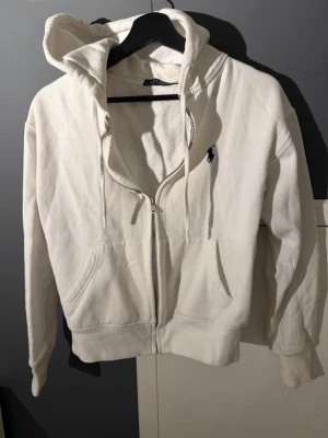 Vit hoodie från Polo Ralph Lauren - Klassisk vit hoodie med dragkedja från Polo Ralph Lauren. Har huva med dragsko, två fickor framtill och den ikoniska mörkblå loggan broderad på bröstet. Tillverkad i mjuk bomullsmix som känns skön mot huden. Perfekt för en avslappnad och stilren look.