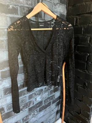 Svart spetstopp  - Säljer en svart, transparent spetsblus från Gina Tricot. Blusen har v-ringning, lång ärm och är croppad i modellen. Perfekt för dig som vill ha en edgy och trendig topp med snygga spetsdetaljer.