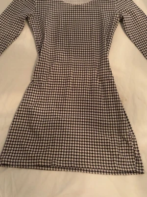 Svartvit mönstrad klänning - Snygg svartvit klänning med klassiskt rutigt mönster. Modellen är figurnära med lång ärm och kort längd. Perfekt för dig som gillar en stilren och trendig look. Materialet känns mjukt och stretchigt, troligen polyester eller bomullsmix.