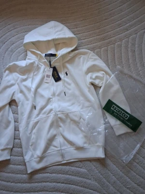 Vit hoodie från Polo Ralph Lauren - Vit hoodie med dragkedja från Polo Ralph Lauren. Klassisk design med huva och dragsko, två fickor framtill och den ikoniska svarta polospelaren broderad på bröstet. Tillverkad i mjukt bomullsmaterial för en skön och avslappnad look.