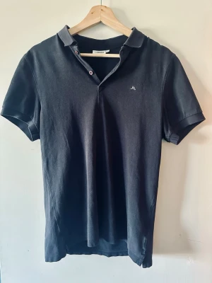 Marinblå pikétröja från J.Lindeberg - Klassisk marinblå pikétröja från J.Lindeberg med liten logga på bröstet. Tröjan har korta ärmar, krage och knappar vid halsen. Perfekt till jeans eller shorts för en clean look.