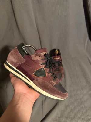 Philippe Model  - Snygga vinröda sneakers från Philippe Model med detaljer i svart och beige. Ovandelen är i mocka och textil, med svarta skosnören och en vit sula med svart rand. Klassisk låg modell med coola kontrastpaneler och retrovibbar.