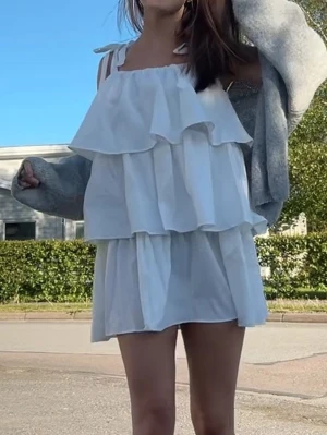 Vit volangklänning med knytband - Söt vit kortklänning med volanger och knytband på axlarna. Klänningen har en luftig passform och är perfekt för varma dagar. Tillverkad i ett lätt bomullsliknande material som känns skönt mot huden. Storlek 36 men passar mig som brukar ha xs-s i klänningar