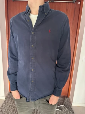 Mörkblå skjorta från Ralph Lauren - Säljer en marinblå långärmad skjorta från Polo Ralph Lauren i Slim Fit-modell (herrstorlek Small). Skjortan har en snygg passform och en broderad logga.  Det finns dock ett litet hål på ena ärmen, vilket framgår tydligt på den sista bilden men syns inte så tydligt när man har på sig den. I övrigt är den i bra begagnat skick. • Märke: Polo Ralph Lauren • Passform: Slim Fit • Storlek: Small (Herr) • Färg: Marinb