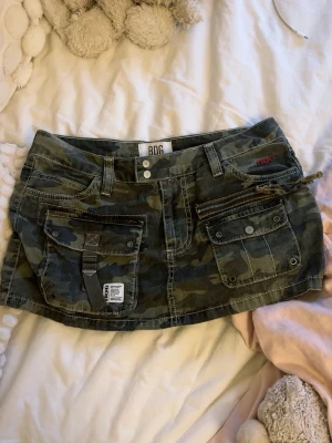 Camouflage kortkjol  - Säljer en grön camouflage kortkjol med coola detaljer som stora fickor, dragkedja och knappstängning fram. Kjolen har bälteshällor och kontrastsömmar för en extra edgy look. Perfekt för dig som gillar streetstyle.
