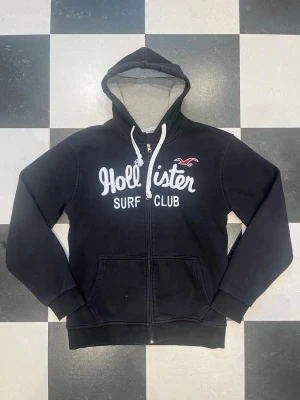 Hollister zip hoodie - Svart hoodie från Hollister med vit broderad text 'Hollister Surf Club' på bröstet och klassisk logga. Dragkedja framtill, två fickor och justerbar huva med vita snören. Står storlek xl men är M. Insidan av huvan är ljusgrå. Perfekt för chill dagar och streetwear vibes.