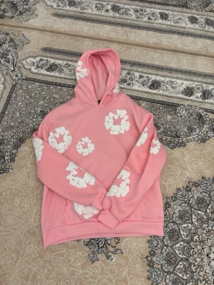 Rosa hoodie med vita blommor - Säljer en rosa hoodie med huva och vita blommönster över hela tröjan. Hoodien har en avslappnad passform och är tillverkad i ett mjukt material som känns skönt mot huden. Perfekt för dig som gillar färgglada och lekfulla plagg.