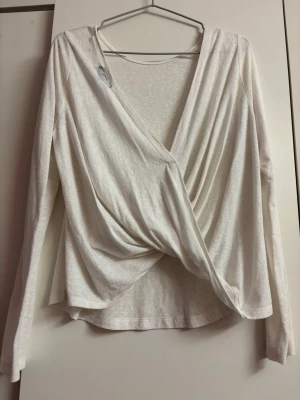 Vit omlottopp med lång ärm - Snygg vit omlottopp med lång ärm och djup v-ringning. Toppen har en draperad front som ger en cool och avslappnad look. Perfekt att styla med jeans eller kjol för en trendig outfit.