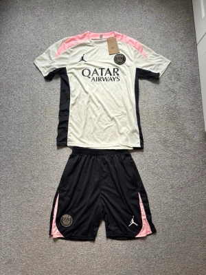 PSG träningsset vit/svart/rosa - Säljer ett PSG träningsset från Jordan i vitt, svart och rosa. Tröjan har Qatar Airways-tryck, PSG-logga och Jordan-logga. Matchande svarta shorts med rosa detaljer och PSG-märke. Perfekt för fotbollsträning eller gymmet.