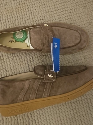Bruna mocka loafers från Adidas - Snygga loafers från Adidas i brun mocka med klassiska detaljer och gummisula. Skorna har diskret Adidas-logga på sidan och ovandelen, samt dekorativa sömmar och en stilren siluett. Perfekta för dig som gillar retrovibbar och streetstyle.