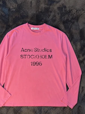 Rosa långärmad topp Acne Studios - Cool rosa långärmad topp från Acne Studios med tryck 'Acne Studios STOCKHOLM 1996' på bröstet. Tillverkad i mjuk bomull med relaxed fit och rund halsringning. Perfekt för dig som gillar streetwear och vill sticka ut lite extra. Storlek M men passar S också